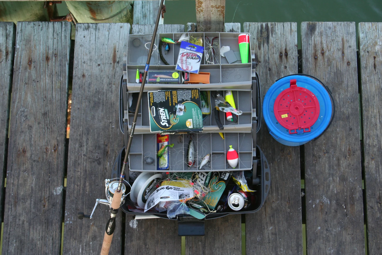 An open tackle box, fishing rod and bait bucket on wooden boards.