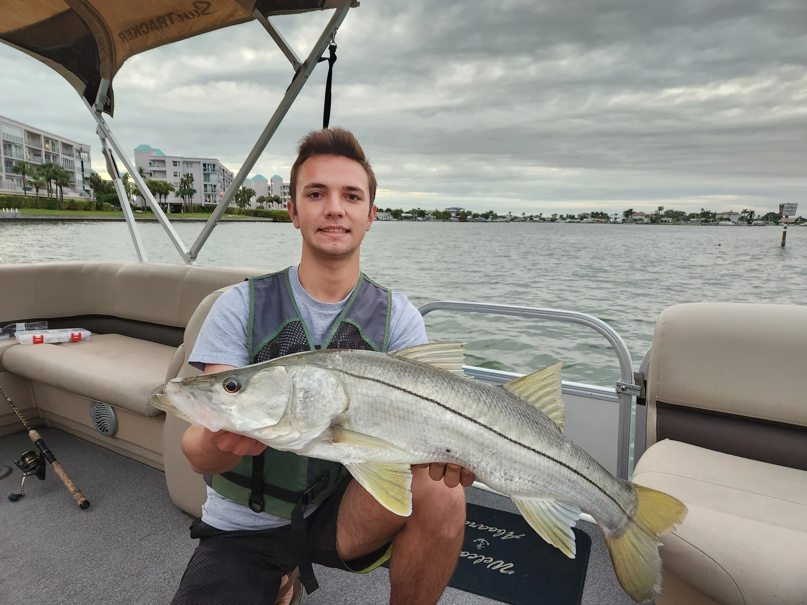 An angler supporting a whole common snook horizontally aboard a boat, with a prominent black lateral line and yellowish fins; catch location unconfirmed.