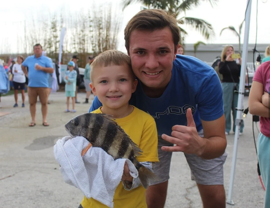 A child holding a sheepshead beside an adult at a waterfront gathering; catch location not recorded.