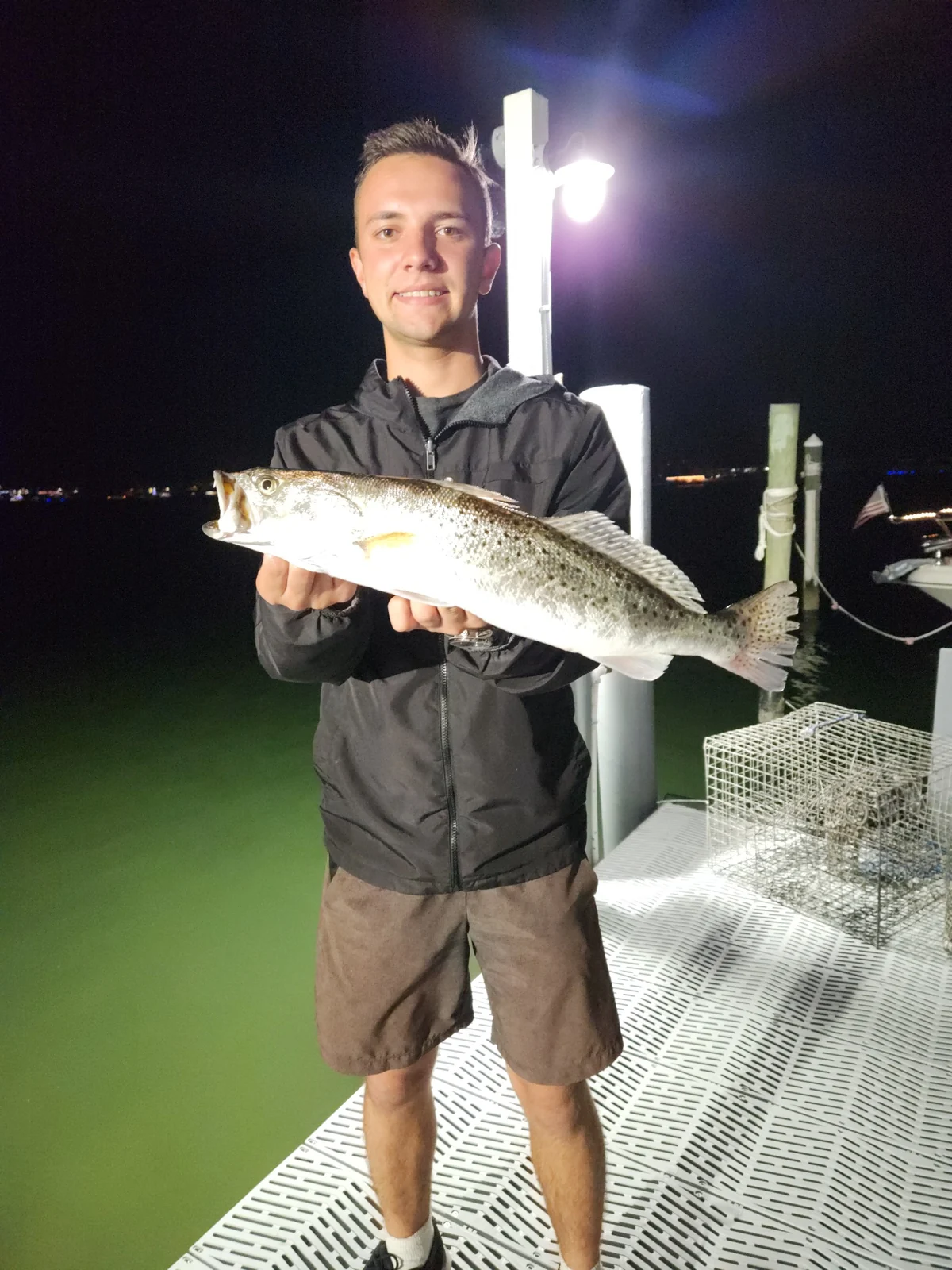 A whole spotted seatrout held horizontally at a dock at night, showing dark spots along its silver body and tail; catch location unconfirmed.