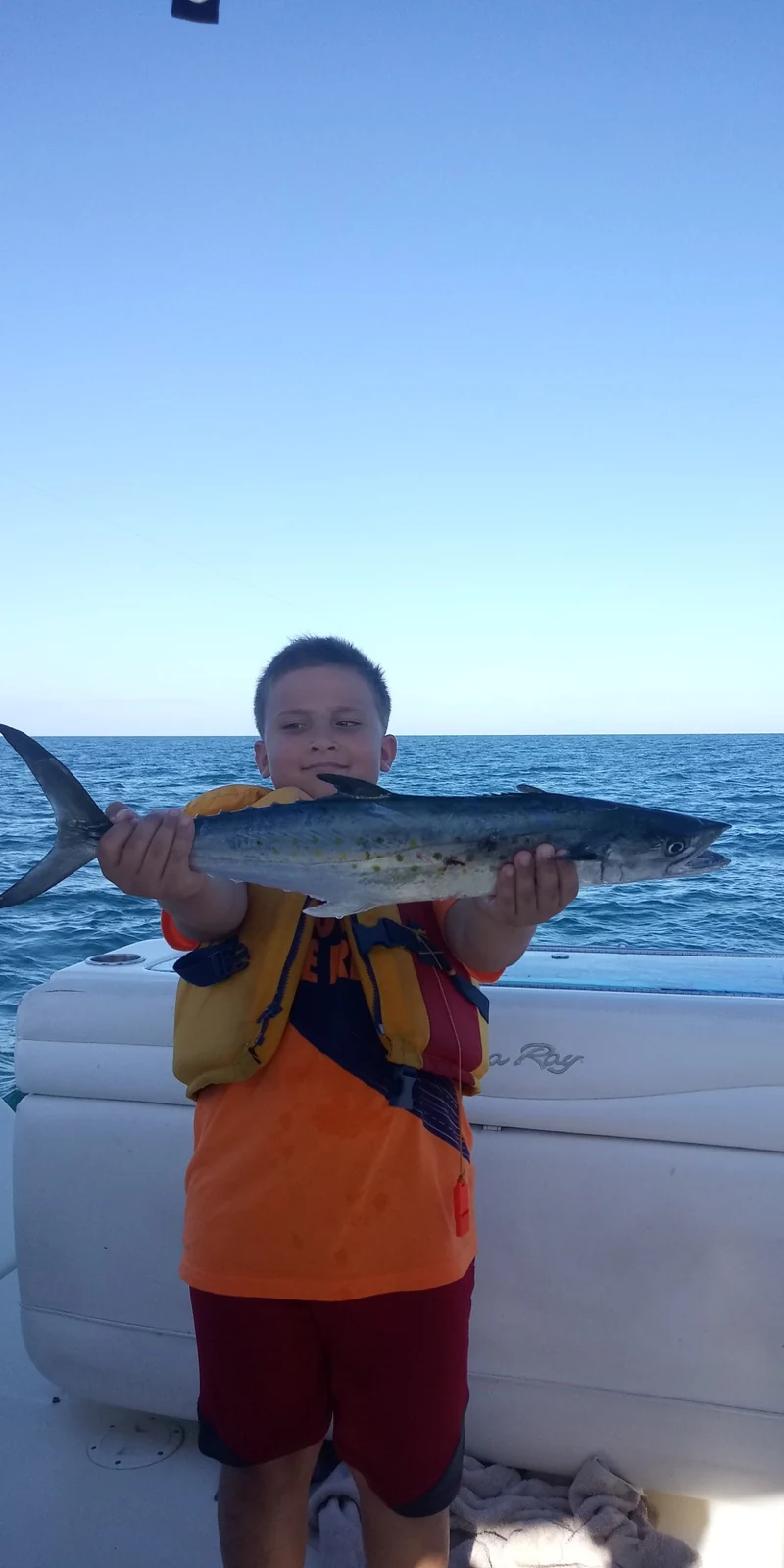 A young angler wearing a life jacket holds a silver fish horizontally aboard a boat; species and location are unconfirmed.